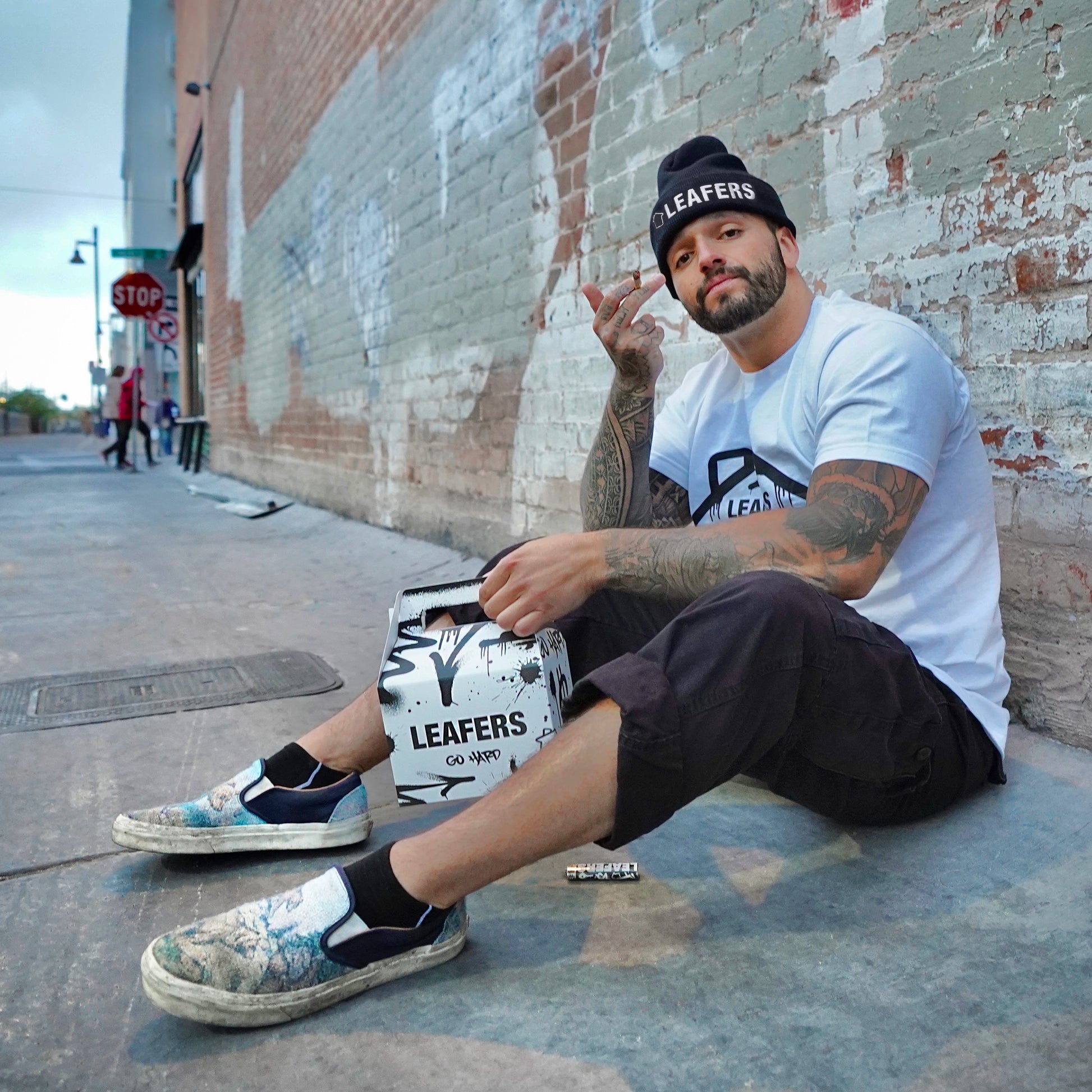 Man sitting on a sidewalk with a 'Leafers' Trappy Meal, wearing a white t-shirt and black shorts.
